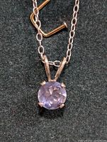 Close-up photo of silver chain necklace with round faceted light purple tanzanite pendant in four-prong silver setting on dark background.