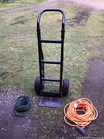 Full view of black Harper hand truck standing upright on grass and gravel, with a green extension cord coiled on the left and an orange extension cord coiled on the right.