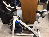 Front and side view of white steel iDeer Life exercise bike with black and silver seat, handlebar and blue-accented flywheel