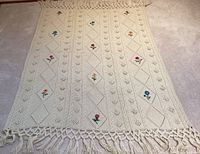 Full view of cream colored knit blanket featuring rows of diamond patterns with embedded embroidered flowers and surrounding cable and bobble knit textures. Fringe detail visible at bottom edge.