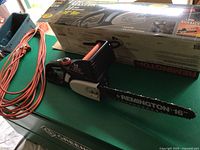 Remington electric chain saw with 16-inch bar next to a long orange extension cord and original packaging box on a green surface.