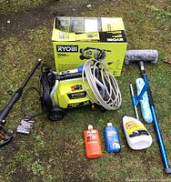 Overall view of Ryobi 1900 PSI electric pressure washer with accessories and original box all laid out on grass.