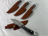 Four fixed blade knives in brown leather-like belt sheaths laid out on white cloth. Three knives unsheathed and one still in sheath, each sealed in plastic wrapping.