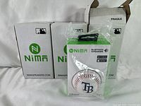 Three Nima branded speaker boxes stacked side by side, one open showing a plastic-wrapped baseball speaker with Tampa Bay Rays logo.
