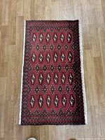 Full view of the small rectangular rug laid flat on a wood floor, showing overall geometric pattern and color scheme in red, brown, black and cream with white fringe edges.