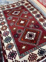 Close-up photo showing the red central field of the Shiraz area rug with its geometric diamond and cross patterns, bordered by a cream band with multicolored medallion motifs.