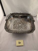 Front view of the square silver plated dish with ornate floral and scroll embossed design and attached handle showing details and patina.