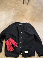 Black Geiger wool sweater laid flat on carpet with Eaton red leather gloves on top of left pocket. Shows full sweater front and gloves.