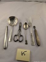 Full view of six sterling silver and steel items including spoons, scissors, fork, and tools laid out on a white cloth.
