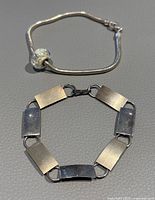 Two 7-inch sterling silver bracelets shown. One with linked rectangular plates, the other a snake chain with large glass charm bead.