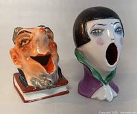 Front view showing two ceramic head figurines with open mouths. Male head on book base, female head with bob hair and purple collar.