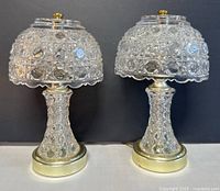 Pair of vintage cut glass table lamps shown from front on a white surface against a dark background. The lamps have clear patterned cut glass shades and bases with brass accents. Both lamps have brass finials on top of the shades.