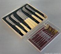 Top view of 7 cheese and butter spreaders with cream and black handles by Hercules Inox in original box alongside 6 ornate teaspoons by ALP Kultakeskus in box