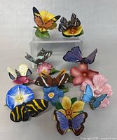 Nine porcelain butterflies displayed on colorful flower bases, showing all the figurines in the lot, the delicate painting and shapes of both butterflies and flowers.
