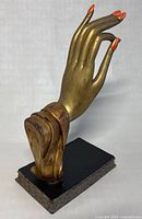 Side profile of mannequin hand showing orange fingernails and gold wood finish, mounted on black decorative base with ornate edging.