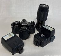 Canon AV-1 film camera with lens, separate telephoto zoom lens, and two camera flashes displayed on a flat surface.