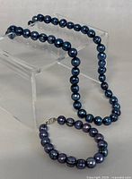 Photo of dark blue pearl necklace and matching bracelet displayed on clear stand for size comparison and style.