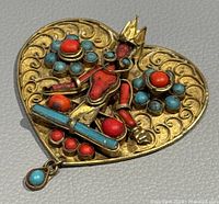 Clear angled close-up of heart-shaped brass brooch featuring Hindu god figure with red coral and turquoise inlay.