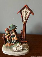 Front side view of the Hummel figurine showing two children sitting on a wooden fence, a crucifix shrine above, and a sleeping dog at the base.
