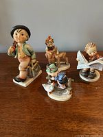 Four collectible Hummel figurines displayed on a wooden surface showing each figurine's front side and pose.