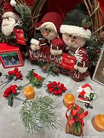 Photo showing three snowman plush Christmas decorations holding a 'Merry Christmas' sign, red berry sprigs, small candles, and a wooden Santa ornament.
