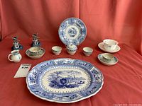 Overview of collection on red cloth background showing all ceramic pieces including two Delft windmills, two large decorative plates, six small bowls, three teacups with saucers, and small covered sugar bowl.
