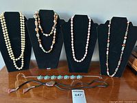 Six necklaces displayed on black necklace stands and flat on surface, showing variety of stone and bead have necklaces including bolo tie.