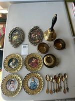 Top view of the brass decorative items including plates with portraits, bell, creamer and sugar containers, ashtray, and spoons arranged on table.