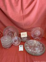 Photo showing all pieces of the clear glass dinnerware collection with floral embossed patterns against a pink fabric background.