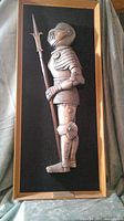 Full frontal image of cast stone armor relief mounted on black suede within wood frame showing side profile of foot soldier holding halberd.