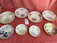 Overview of all 8 plates with varied floral hand-painted designs and scalloped edges on a red cloth backdrop.