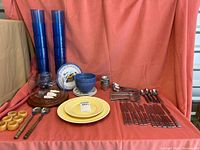 Wide view showing buttercream ceramic plates, Cambridge flatware, blue plastic tumblers, bowls, and other serving accessories on a red cloth backdrop.