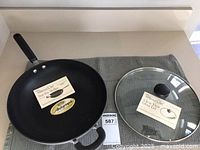 Both the black 12 inch stir-fry skillet and the clear glass lid shown on a surface with product labels attached, new in box.