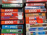Photo showing multiple stacked 1000 piece puzzle boxes predominantly by Guild with other brands visible including Big Ben and MB