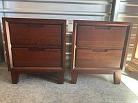 Front view of two dark wood bedside tables side by side showing two drawers each with recessed handles and solid legs.