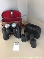 Both pairs of binoculars with their respective leather cases placed side by side showing size difference and overall condition.