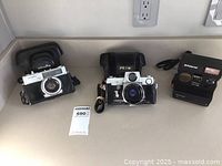 Front view of three vintage cameras placed side by side on countertop: Minolta 7S, Petri Flex 7, Polaroid Sun 660 instant camera.