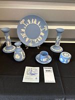 Full set including plate, pair of candle holders, vase, demitasse cup and saucer, keepsake box, and informational booklet displayed on black cloth.