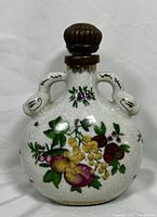 Front view of the white crackle porcelain moon flask with colorful hand-painted fruit and floral decoration, gold snake-shaped handles, and brown metal stopper on top.