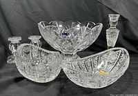 Mixed collection of clear lead crystal items displayed together on black background: frosted candlestick, footed bowl with sticker, cut crystal bowls, and candle holders