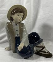 Front and side view of the porcelain figurine showing the boy seated with detailed clothing and hat, glossy finish, and facial features.