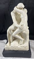 Front view of white plaster sculpture of two nude figures in intimate embrace on black painted base.