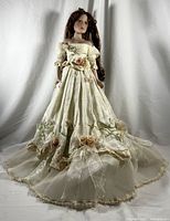 Full view of the porcelain doll in off-white floral dress standing against white fabric background showing full length and elegance.
