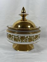 Front view of the ceramic jar showing white body with gold floral pattern, pedestal base, and domed gold lid with pointed finial