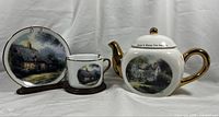 Front view of Thomas Kinkade teapot, teacup and saucer showing cottage artwork and gold trim. Teapot lid and pot have cracks.