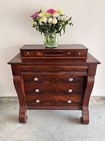 Front view of antique Empire bureau showing polished wood surface, three large drawers, upper section with smaller drawers, and bouquet of flowers on top.