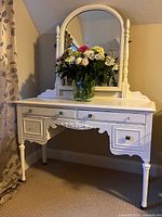 Front view of white antique wooden vanity with arched tilting mirror, floral arrangement on top, turned legs, and storage drawers.