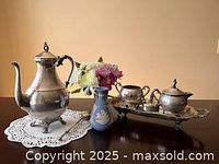 Silver plated tea set with teapot, sugar bowl, creamer, and blue Wedgwood vase placed on a dark wood table with lace doily underneath teapot.