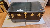 Closed black antique steamer trunk with brass locks and studs, placed on floor.