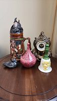 Photo showing all items in the lot arranged on a wooden table: stein, clock, bell, decanter, and duck figurine.